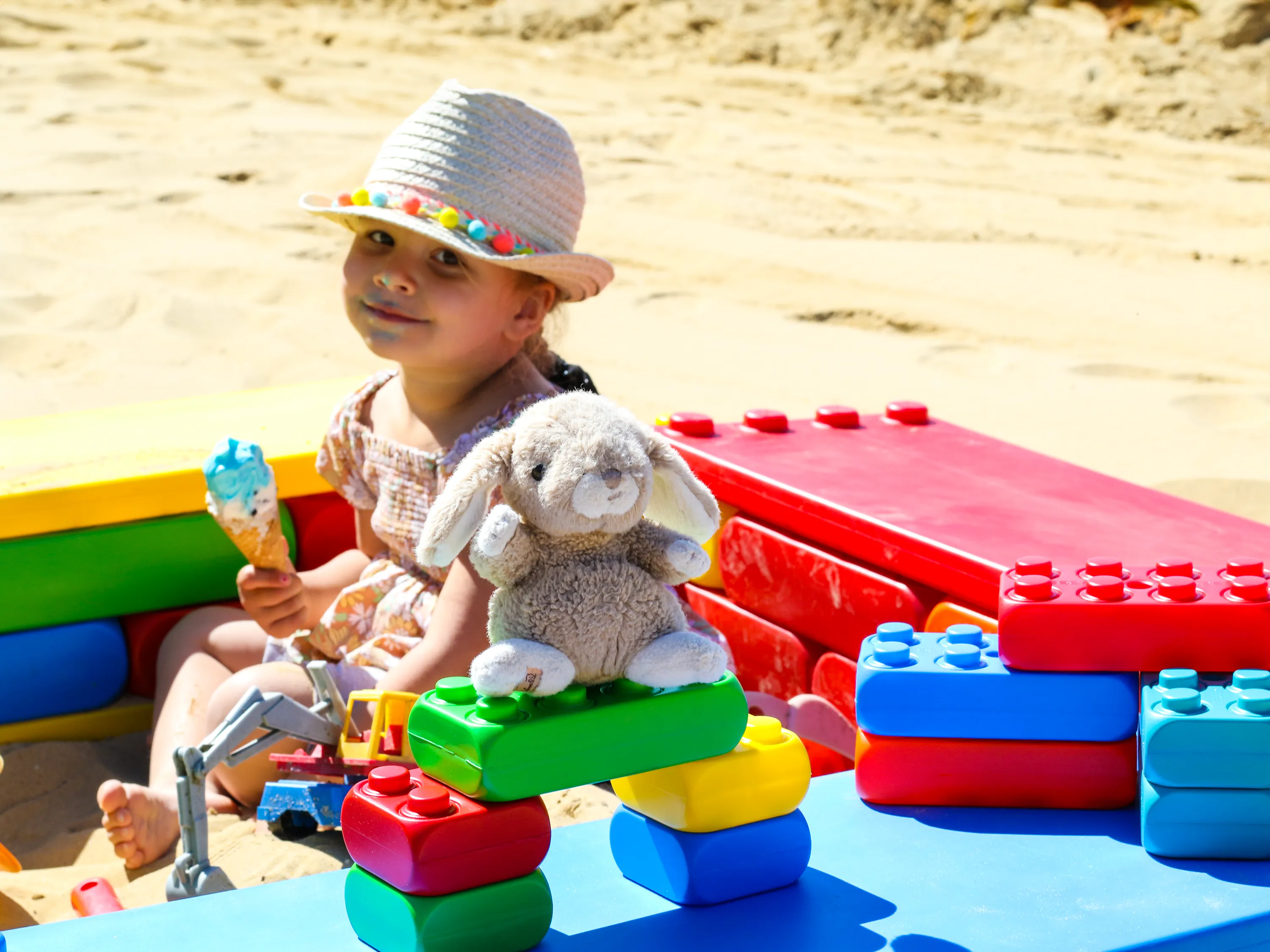 Smiling child wearing a sun hat, sitting in a sandbox made of large, colourful building blocks, holding an ice cream, with a plush bunny placed on the blocks.