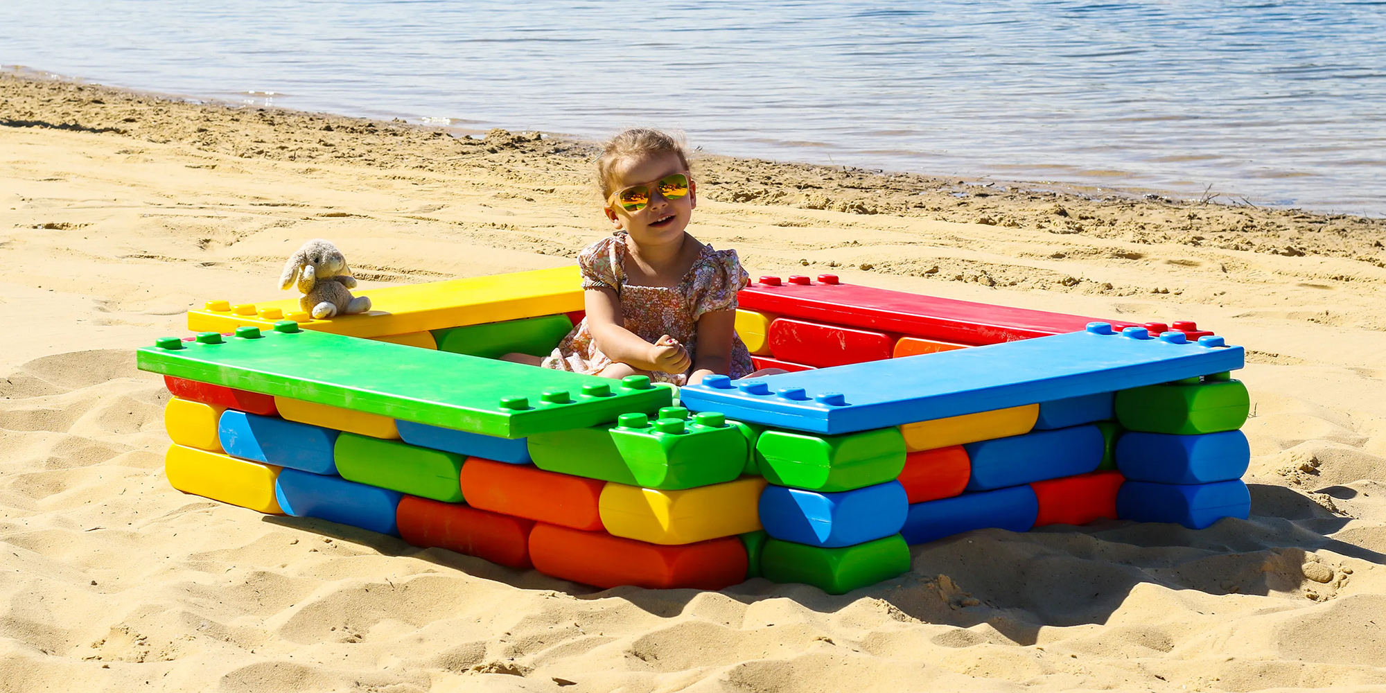 Child playing in a sandbox made of large, colourful building blocks on a sandy beach near the water.