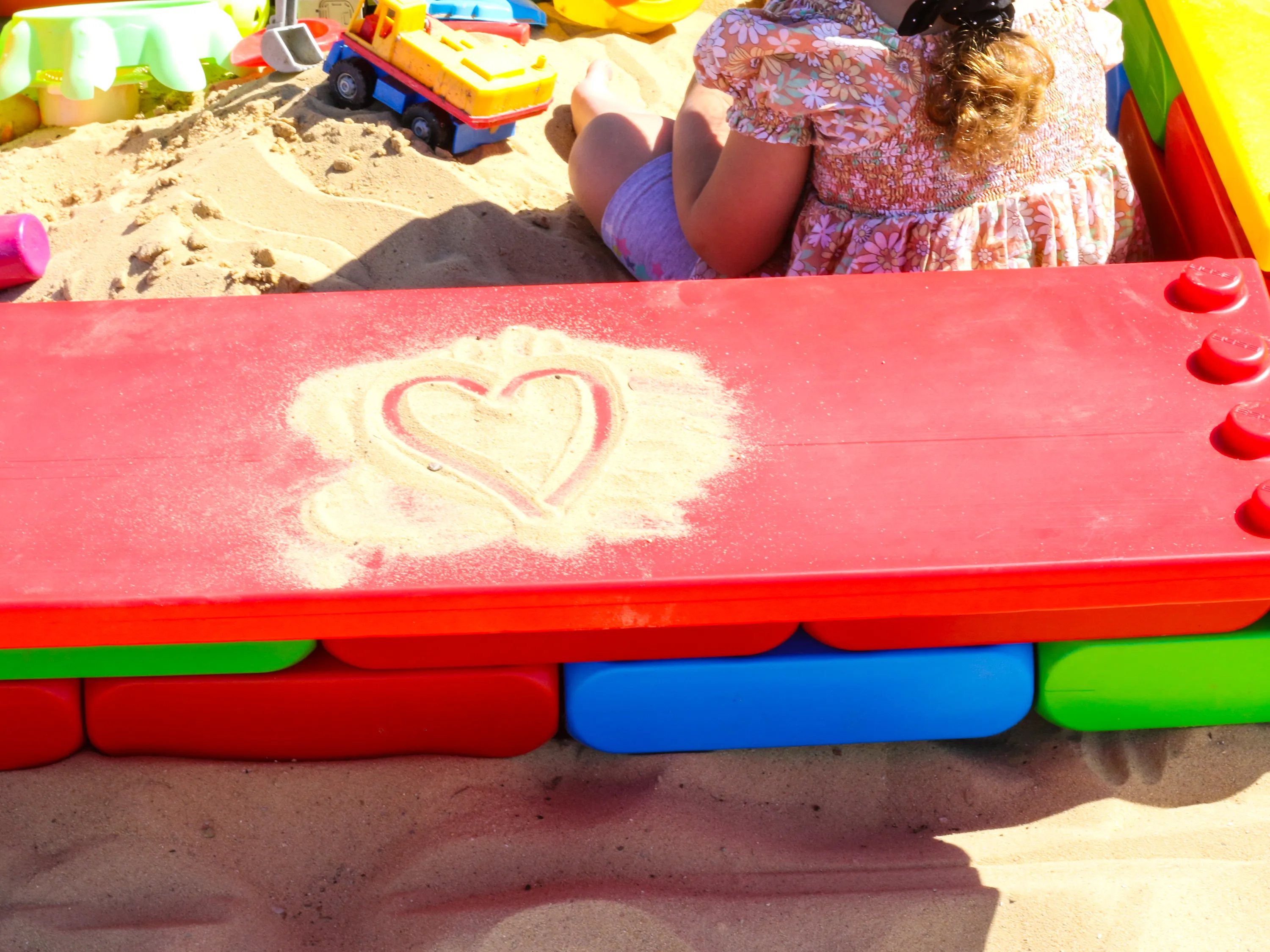 Close-up of a red building block bench on a sandbox with a heart drawn in the sand, while a child plays inside the sandbox.