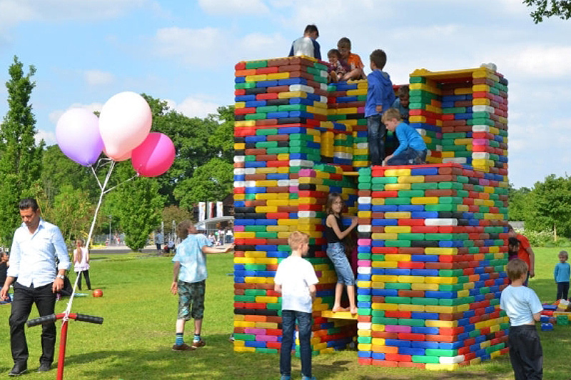 Children playing on a large castle-like structure built from colourful building blocks in a park, surrounded by adults and balloons.