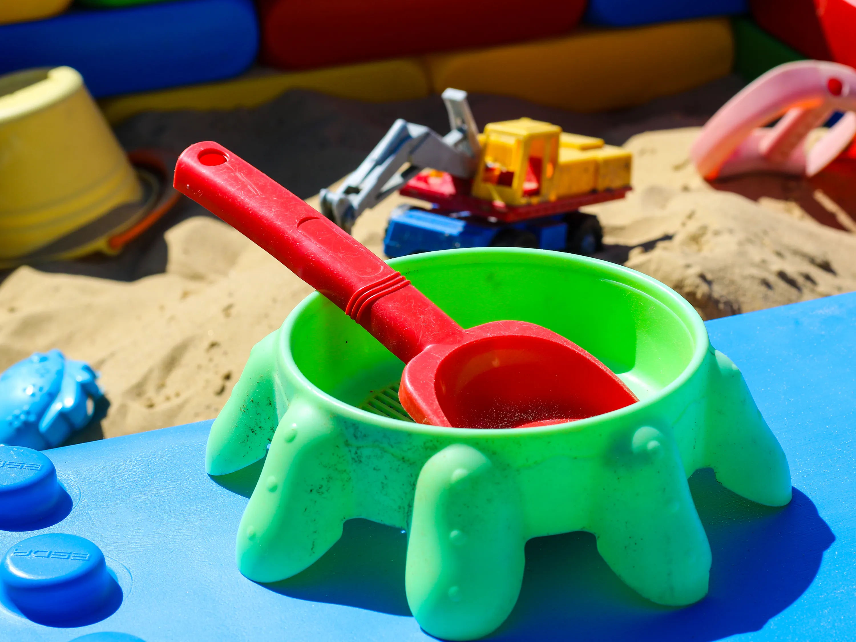 Close-up of a green sand mold and a red shovel on a blue building block, with various sand toys scattered in the background.