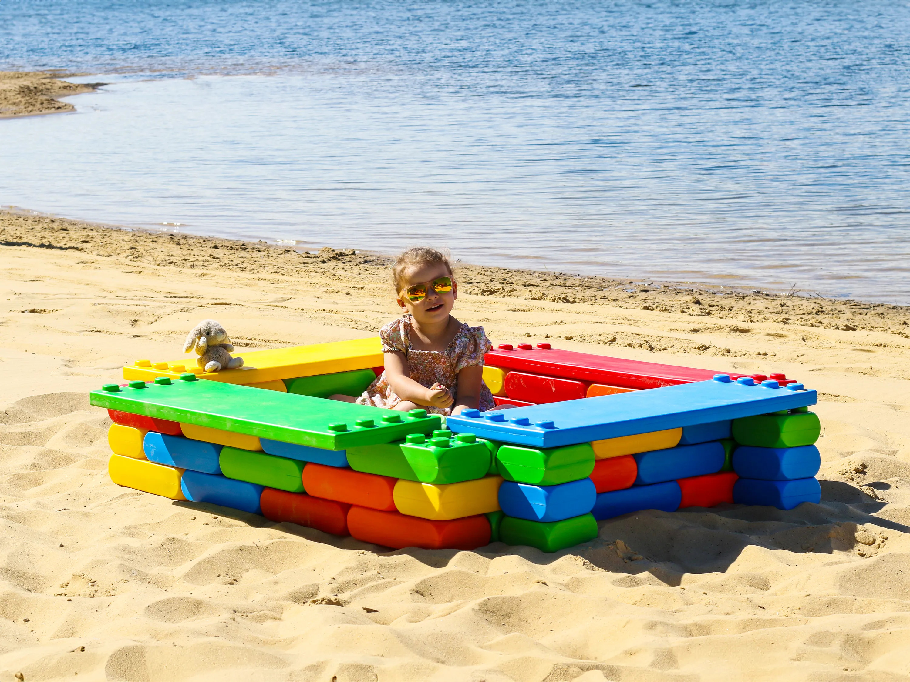 Child playing in a sandbox made of large, colourful building blocks on a sandy beach near the water.