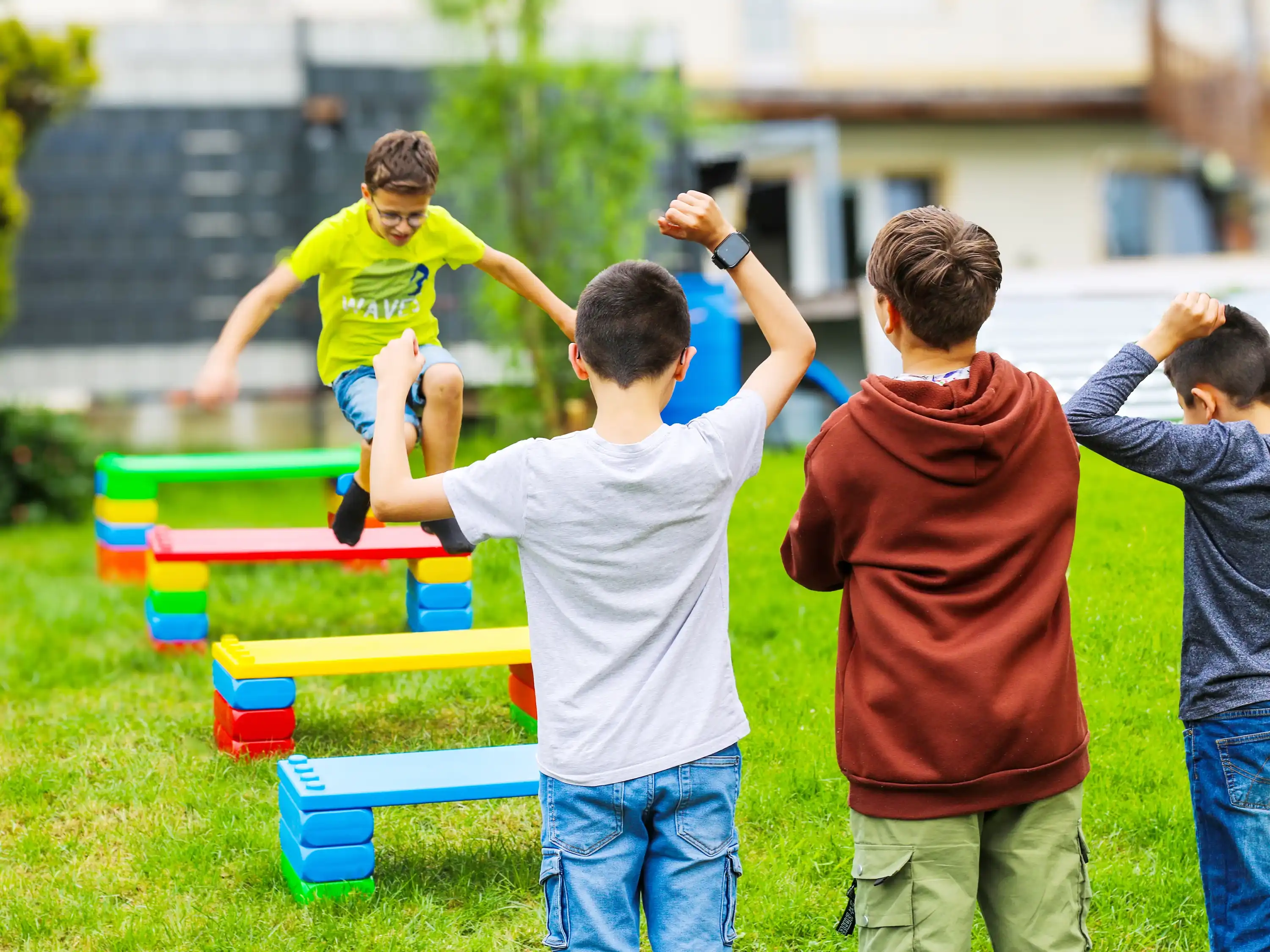 image Children playing outdoors, cheering as one child jumps over colourful benches made of large building blocks on a green lawn.