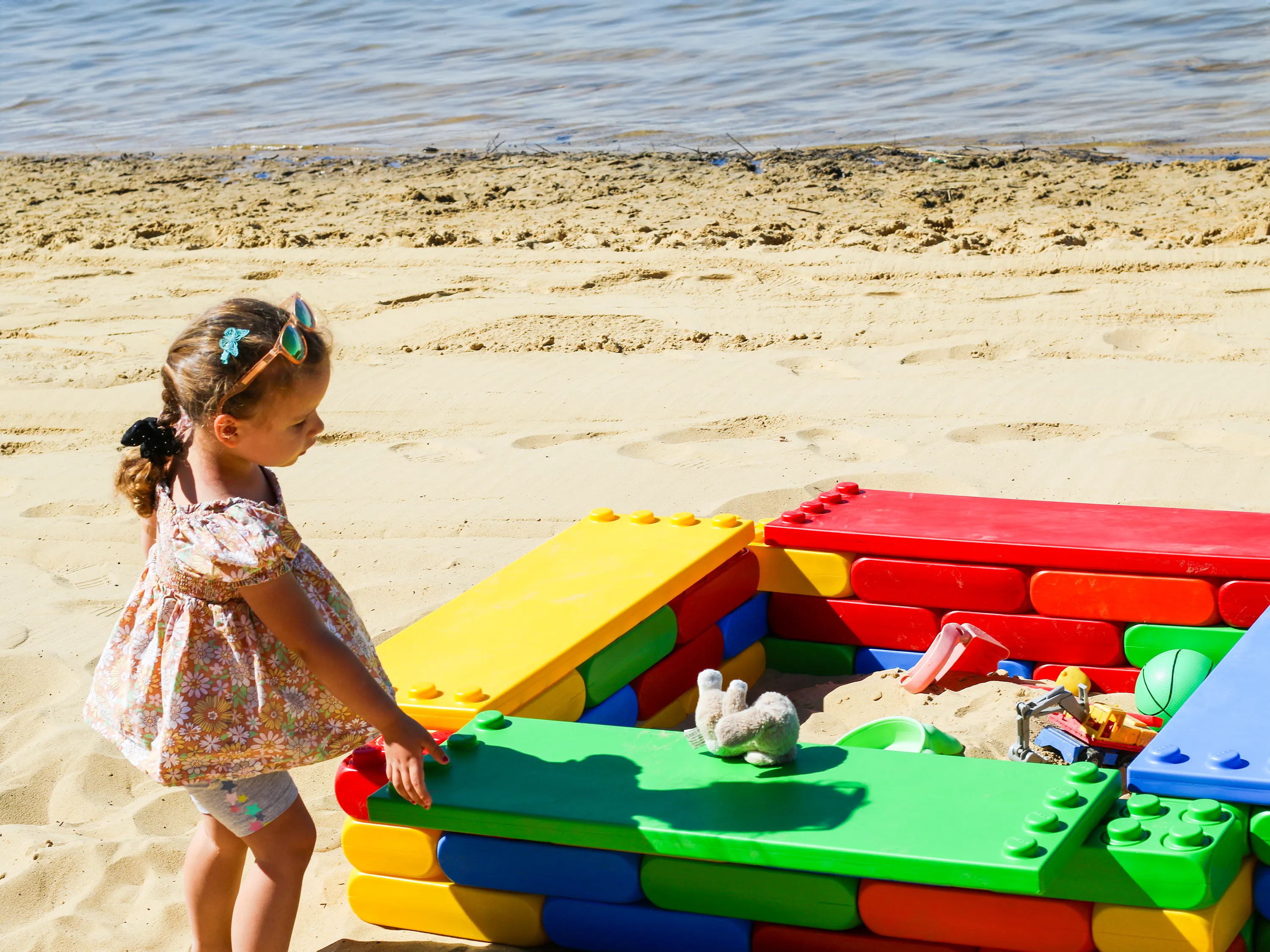 Young girl playing near a sandbox made of large, colourful building blocks on a sandy beach by the water.